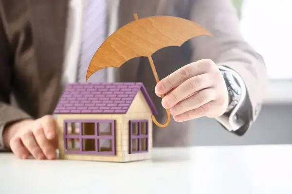 Main d'un homme tenant un parapluie en bois au-dessus d'une maison miniature. Une image symbolisant l'assurance habitation obligatoire.