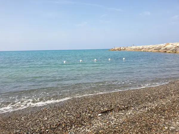Plage de galets à Cecina Mare, avec des bouées blanches marquant la zone de baignade et une jetée rocheuse au loin.