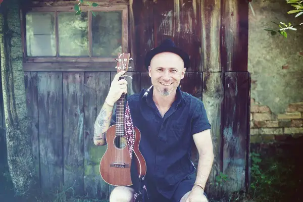 Uśmiechnięty mężczyzna w kapeluszu trzyma ukulele, gotowy do grania nim zajdzie słońce.