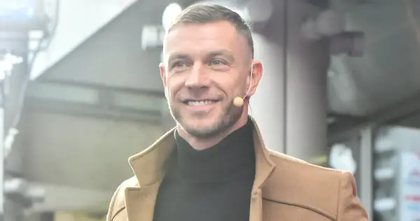 Uśmiechnięty Dariusz Pachut w beżowym płaszczu i czarnym golfie, z mikrofonem przy uchu. Może opowiada o swojej żonie.