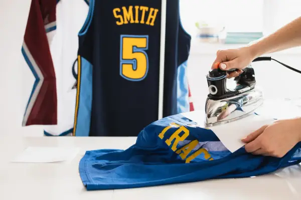 Une main utilise un fer à repasser pour défroisser un maillot de basket bleu avec des lettres jaunes. D'autres maillots sont suspendus en arrière-plan.