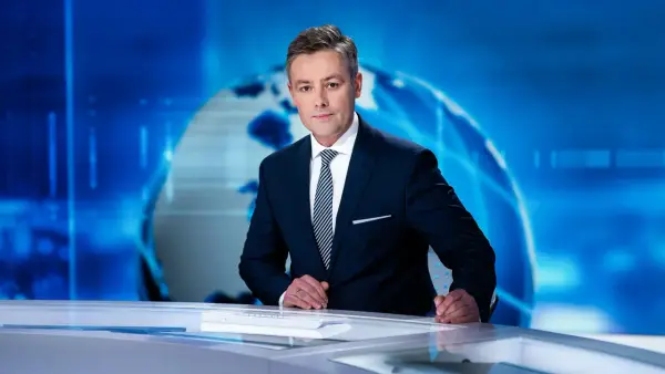 Elegancki konferansjer w garniturze stoi przy biurku na tle niebieskiej grafiki z globusem.