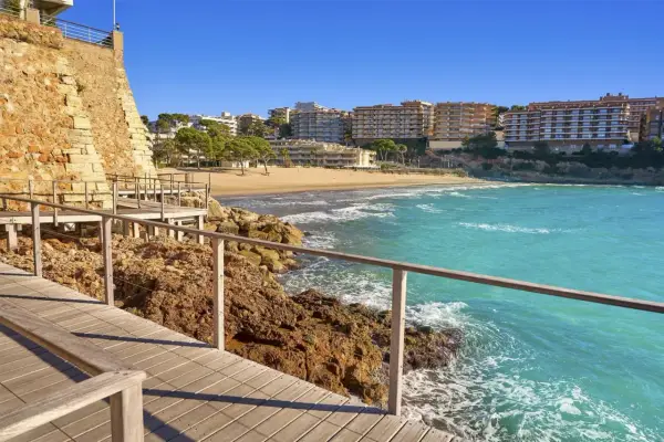 Paseo marítimo de madera junto a rocas y mar turquesa, con playa y edificios al fondo. Un tramo del camí de ronda Tarragona.