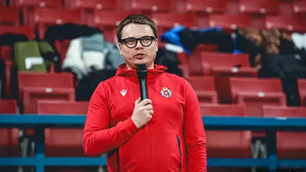 Właściciel Wisły Kraków w czerwonej bluzie z kapturem i mikrofonem, na tle pustych czerwonych trybun stadionu.