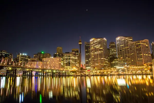 Sydney nocą, z mostem i odbijającymi się w wodzie światłami. Zastanawiasz się, ile się leci do Australii, patrząc na ten widok?
