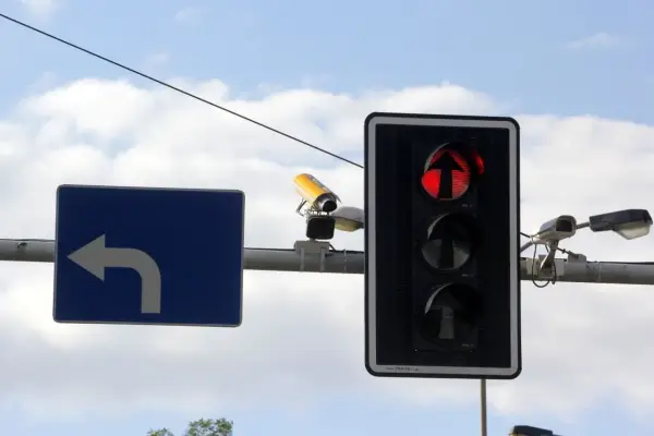Rejestrator przejazdu na czerwonym świetle, widoczny na tle nieba, wraz z sygnalizacją świetlną i znakiem nakazu skrętu w lewo.