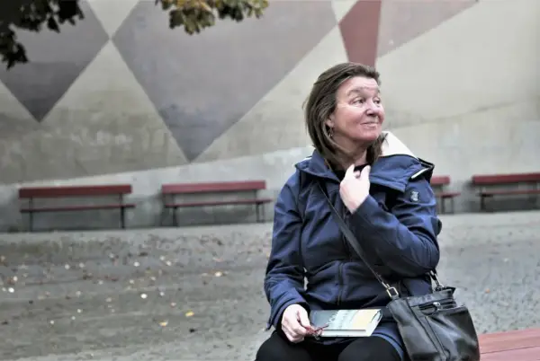 Elisabeth Rynell sitter på en bänk med en bok i knät, blickar ut mot en geometrisk vägg.