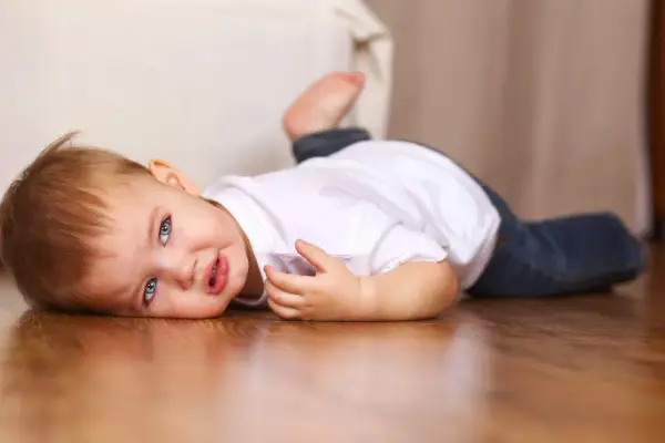 Un bebé de 16 meses con ojos azules y cabello castaño yace en el suelo de madera, vestido con una camiseta blanca y jeans.