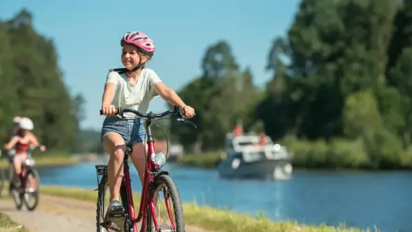 Glad flicka på cykel med hjälm njuter av en cykelsemester med barn längs en kanal. En båt syns i bakgrunden.