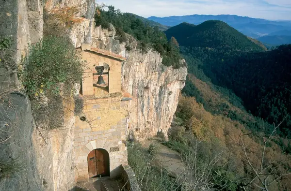Ermita en la costa pubilla, aferrada a un acantilado rocoso con vistas a un valle boscoso. Una campana cuelga sobre la entrada.