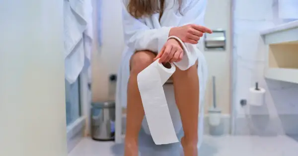 Kobieta w szlafroku siedzi na toalecie, trzymając rolkę papieru. Zastanawia się, czy jej pieniący się mocz to powód do niepokoju.