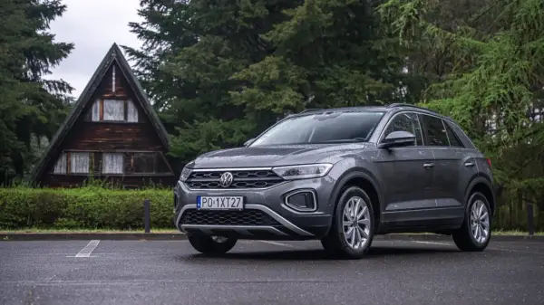 Szary Volkswagen T-Roc używany, zaparkowany przed drewnianym domkiem w lesie. Idealny na wypady za miasto.