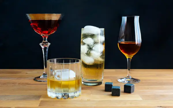 Vier Gläser mit verschiedenen Getränken, darunter ein Glas mit peated whisky, Eiswürfel und Whiskysteine auf Holztisch.
