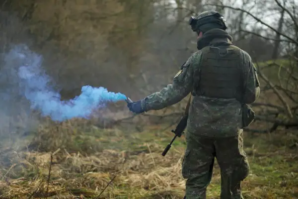 Żołnierz w kamuflażu odpala niebieski granat dymny, przestrzegając zasad ASG.