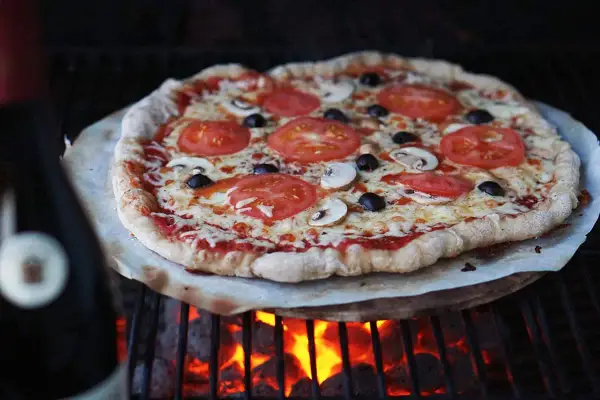 En hemmagjord pizza med tomater, champinjoner och oliver grillas över glödande kol. Perfekt för ett italiensk pizzadeg recept.