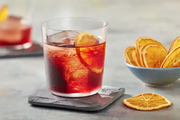 Klasyczny negroni przepis: czerwony koktajl z lodem i plasterkiem pomarańczy, obok miseczka suszonych pomarańczy.