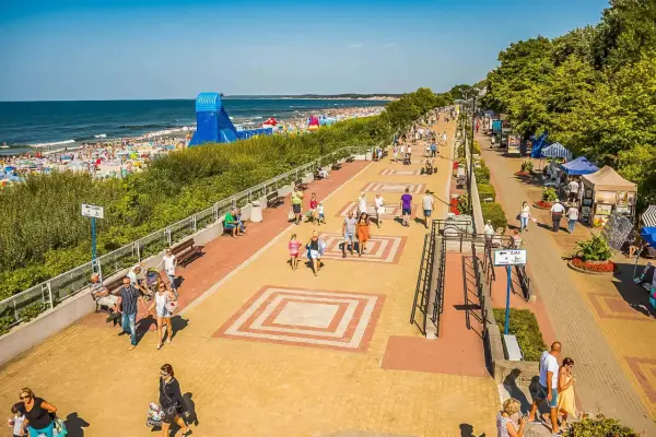 Tętniąca życiem ustka promenada w słoneczny dzień. Ludzie spacerują, odpoczywają na ławkach, a w tle widać plażę pełną turystów i morze.