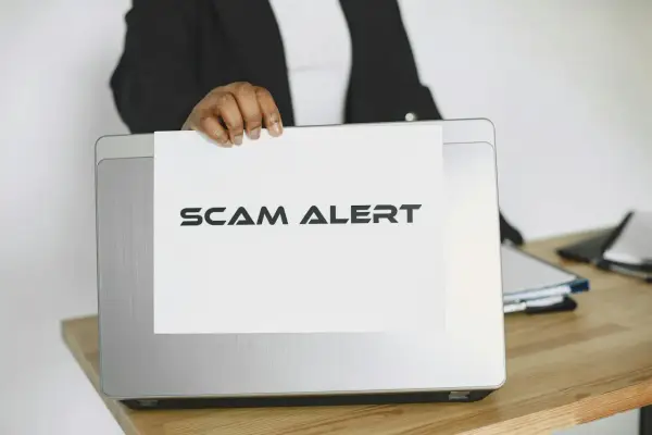 Ostrzeżenie przed oszustwem na Vinted. Ktoś trzyma kartkę z napisem "SCAM ALERT" nad laptopem.
