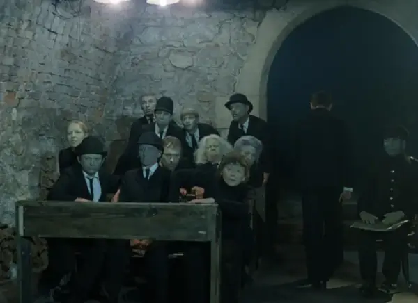 Postacie z "Umarłej klasy" Tadeusza Kantora, w tym uczniowie i nauczyciele, siedzą w ponurej scenerii.