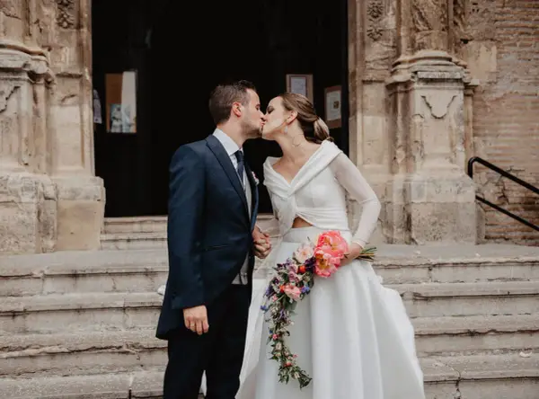 Novios besándose frente a una iglesia, ella lleva un ramo de flores. Un momento especial tras las lecturas de boda religiosa.