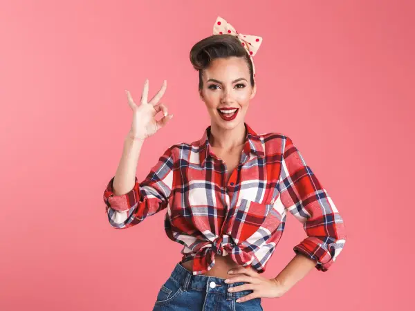 Uśmiechnięta kobieta w stylu pin up girl stylizacje, w kratę koszuli i dżinsach, pokazuje znak OK.