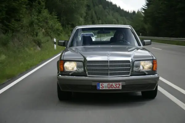 Szary Mercedes W126 jedzie drogą wśród zielonych lasów.