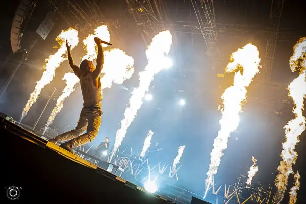 Un chanteur de groupe hard rock des années 2000, micro en main, saute sur scène entouré de jets de flammes spectaculaires.