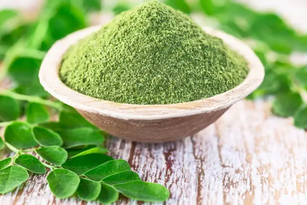 Moringa w proszku w drewnianej misce, otoczona świeżymi liśćmi. Zanim zaczniesz stosować, sprawdź moringa przeciwwskazania.