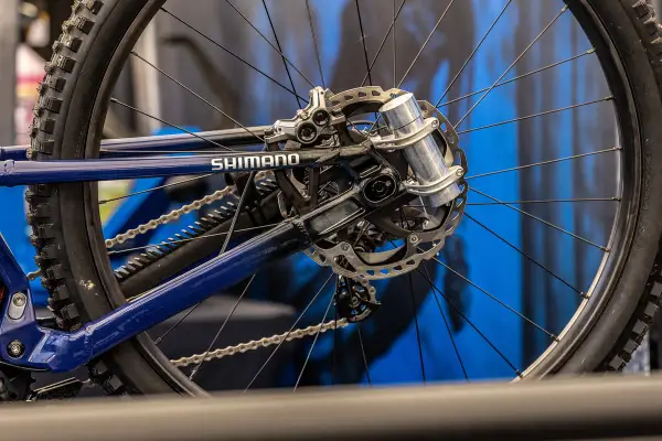 Close-up of a blue bicycle's rear wheel and Shimano drivetrain. A silver grip damper is visible on the axle, ready to absorb shocks.