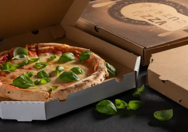 Pyszna pizza w otwartym opakowaniu na pizze, ozdobiona świeżą bazylią. Obok zamknięte kartony.