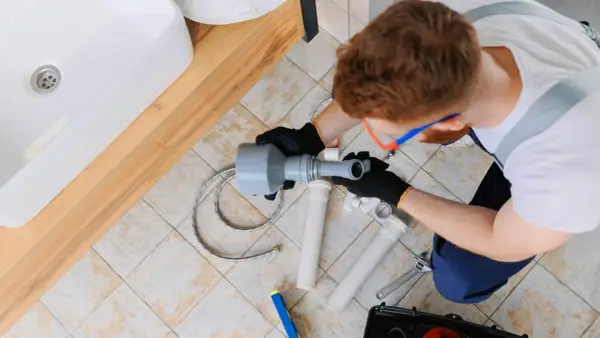 Hydraulik naprawia odpływ, prawdopodobnie z powodu braku odpowietrzenia pionu. Widać umywalkę, narzędzia i elementy instalacji.