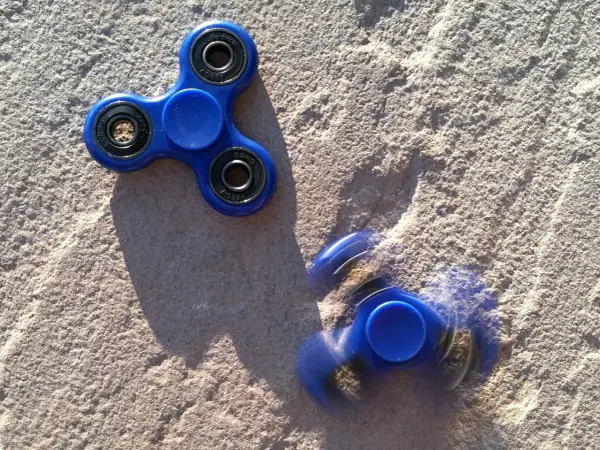 Dwa niebieskie fidget spinnery na piasku, jeden nieruchomy, drugi w ruchu, przypominają czasy, kiedy fidget spinnery były popularne.