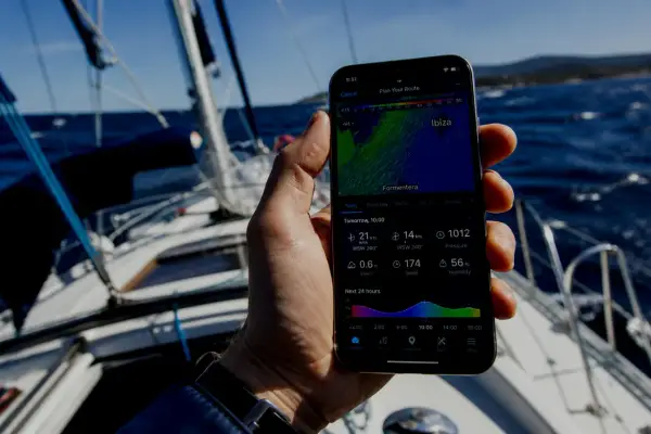 Sur un voilier, une main tient un smartphone affichant une application météo marine gratuite avec les prévisions pour Ibiza.