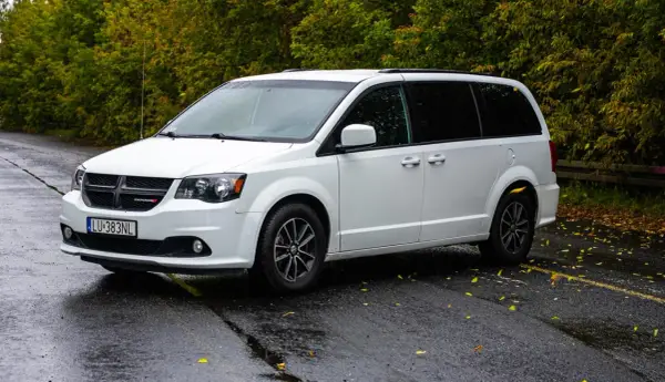 Biały Dodge Grand Caravan na mokrej drodze, otoczony jesienną zielenią.