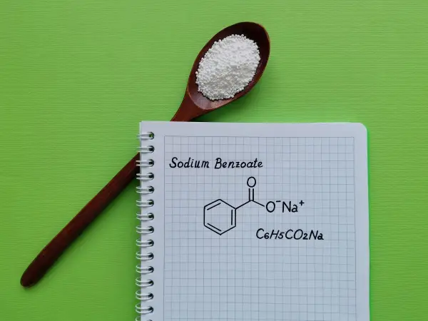 Drewniana łyżka z białym proszkiem, obok notatnik z napisem "Sodium Benzoate" i jego wzorem chemicznym.