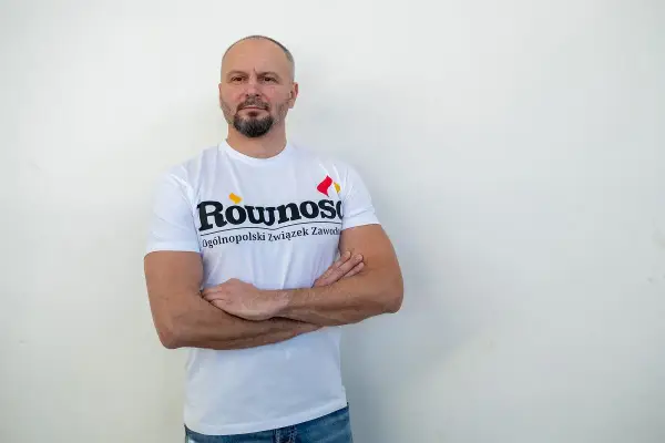 Mężczyzna w koszulce z napisem "Równość Ogólnopolski związek zawodowy" stoi z rękami skrzyżowanymi na piersi.