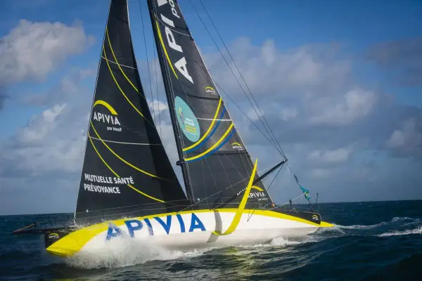 Le voilier Apivia, skippé par Alex Thomson, fend les vagues sous un ciel bleu et nuageux.