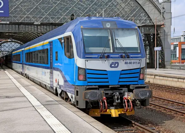 Niebiesko-biały pociąg z żółtym pasem, gotowy do podróży do Czech, stoi na peronie pod nowoczesnym zadaszeniem dworca.