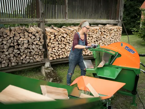 Frau arbeitet mit einer grünen und orangen Wippsäge, um Holzscheite zu schneiden. Im Hintergrund sind Holzstapel zu sehen.