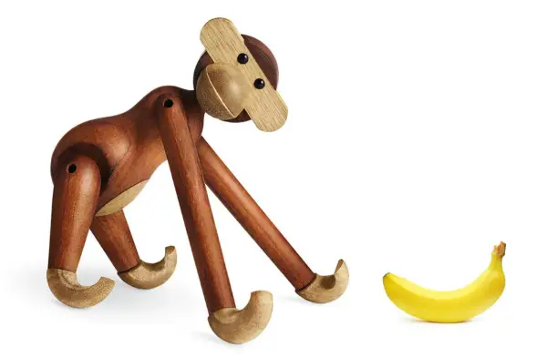 En träapa från Kay Bojesen tittar nyfiket på en banan. Olika kay bojesen apa storlekar finns tillgängliga.