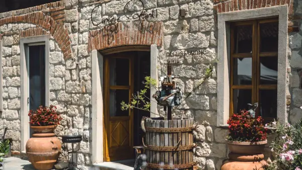Ingresso di "Cinque Quinti", un negozio di degustazione con un torchio antico e vasi di fiori rossi.