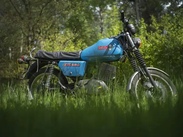 Niebieski motocykl MZ ETZ 250 stojący na trawie, z drzewami w tle.