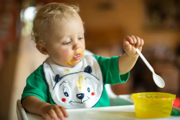 Małe dziecko z marchewkowym puree na buzi i śliniaczku próbuje samodzielnie jeść. Oto jak sprać marchewkę z ubranka!