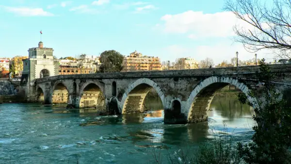 Il **puente Milvio** attraversa il Tevere con i suoi archi antichi, mentre sullo sfondo si stagliano palazzi romani sotto un cielo azzurro.