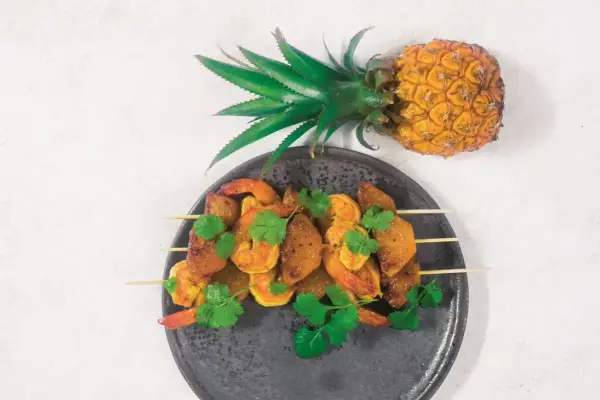 Szpadki z krewetkami i kawałkami ananasa, ozdobione kolendrą. Obok leży cały ananas, symbolizujący owoce egzotyczne.