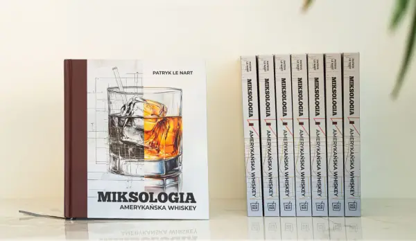 Książka "Miksologia Amerykańska Whiskey" Patryka Le Narta, z ilustracją szklanki z lodem i bursztynowym trunkiem. Obok niej rząd identycznych tomów.