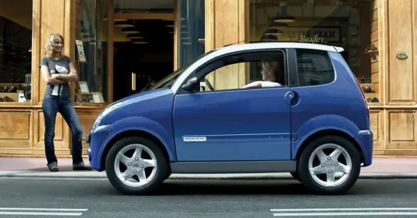 Niebieskie auto od 14 lat, z kobietą za kierownicą, stoi przed sklepem. Obok niego stoi druga kobieta.