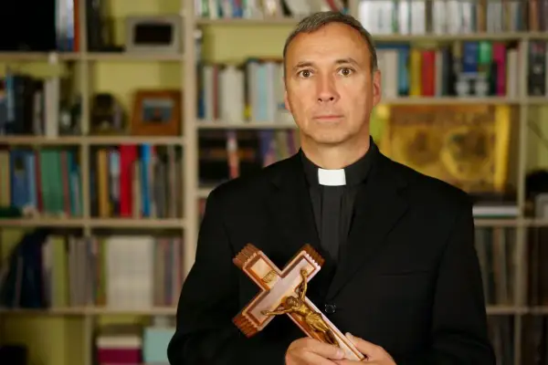 Duchowny z krzyżem w dłoniach, na tle regałów z książkami. Czy religia wlicza się do czerwonego paska?