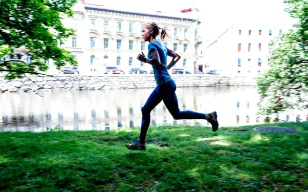 En kvinna joggar långsamt eller går snabbt längs en gräsmatta vid en flod, med byggnader i bakgrunden.