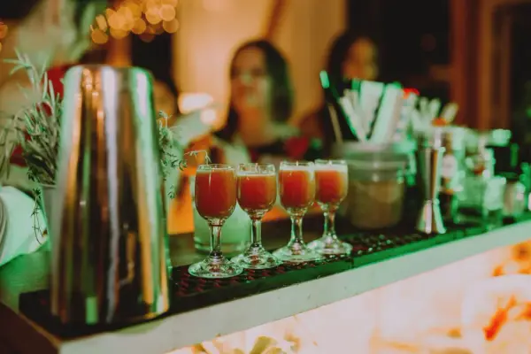 Cztery kieliszki z czerwonym drinkiem stoją na barze, gotowe do podania na weselu. W tle widać gości i dekoracje.